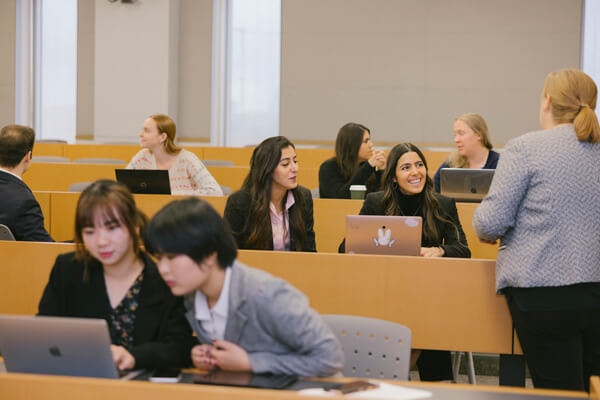 students are seated throughout auditorium style classroom, professor speaks to two students