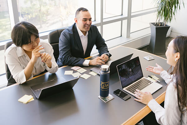 three GW Business students sit around table talking and laughing, laptops open in front of them
