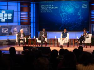 Four people sit on a stage indoors, in chairs in front of an audience. There are monitors that display text in the background and the George Washington University logo. 