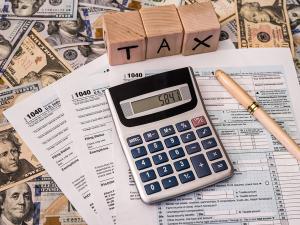 Image shows a desk with a calculator, tax forms, American currency, a pen, and a set of lettered wooden blocks that spell out the word "tax."