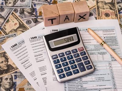 Image shows a desk with a calculator, tax forms, American currency, a pen, and a set of lettered wooden blocks that spell out the word "tax."