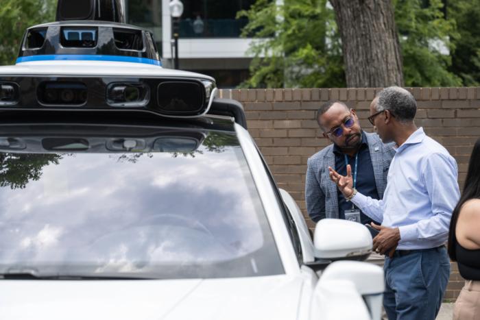 Two people stand next to an autonomous vehicle speaking with one another.