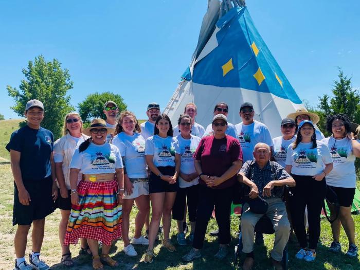 A group of approximately 20 people stand and pose for a photo outdoors. They wear matching shirts that read "South Dakota Native Tourism Alliance." The landscape is has grass and trees. A tepee can be seen behind the group.