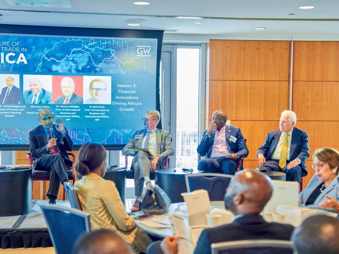 Four people sit on a stage. They wear business attire. One is speaking with a microphone . An audience watches them. A projector displays the word Africa and images of the speakers.