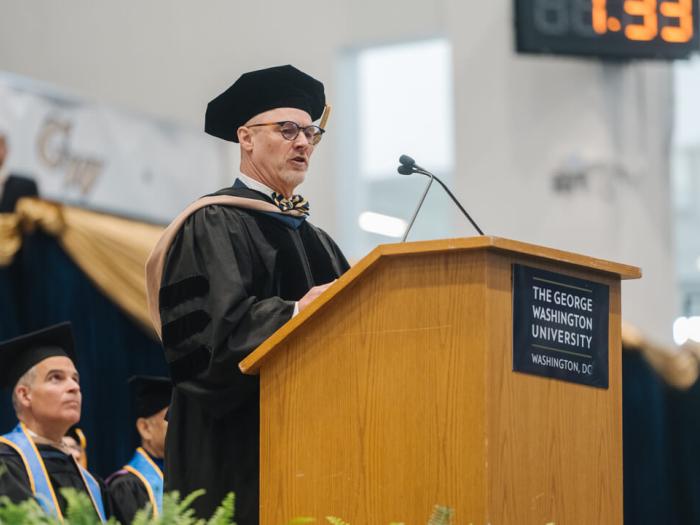 John Roberts, an alumnus of the GW School of Business speaks in front of a podium. He wears regalia.  Others can be seen  on the platform behind him. 