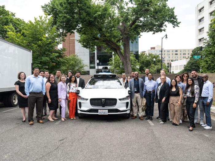 A large group of individuals dressed in professional attire stand in front of an autonomous vehicle. 