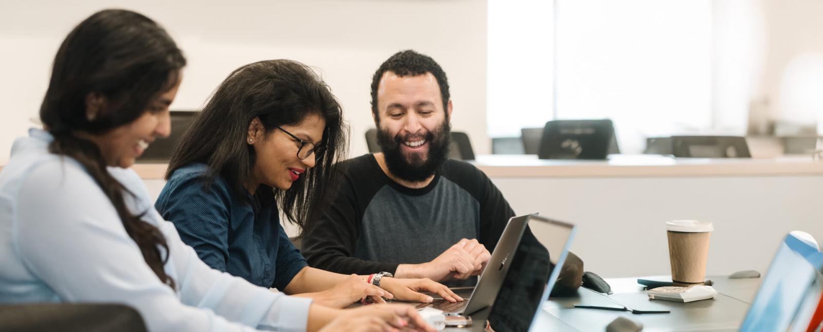 three business students in classroom working on laptops, smiling and talking amongst themselves