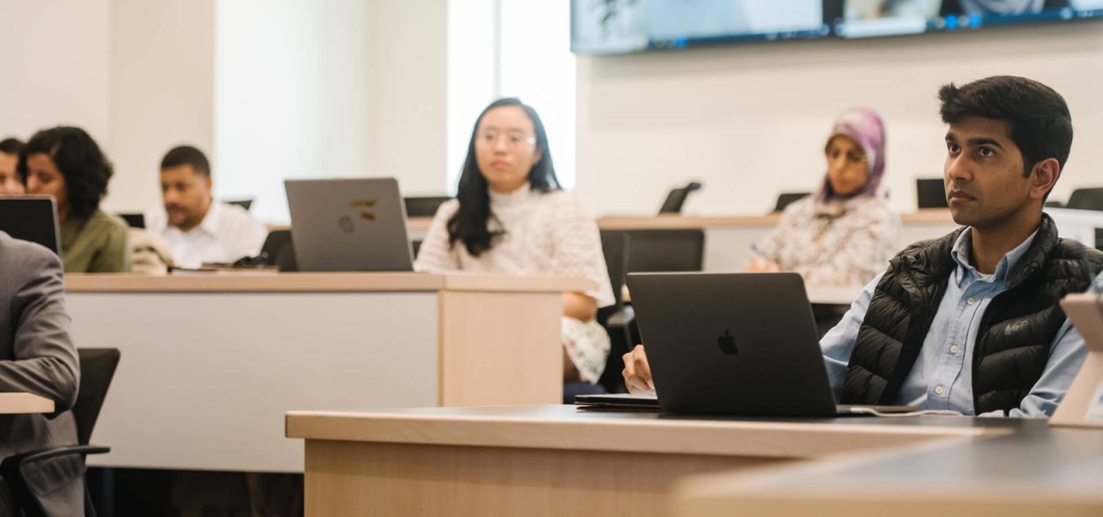 students in classroom listening intently, laptops open in front of them