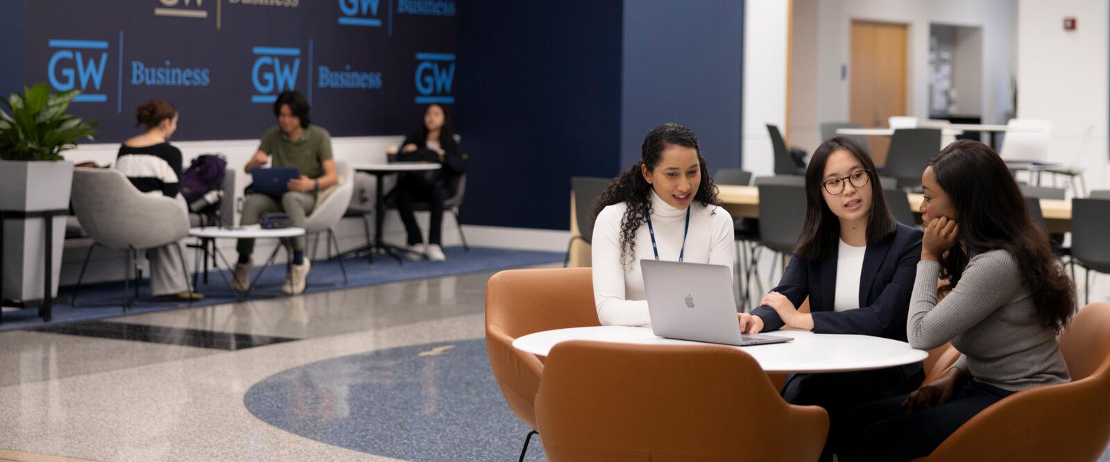 a group of three business students sit around small table in business school lobby, laptop open in front of one
