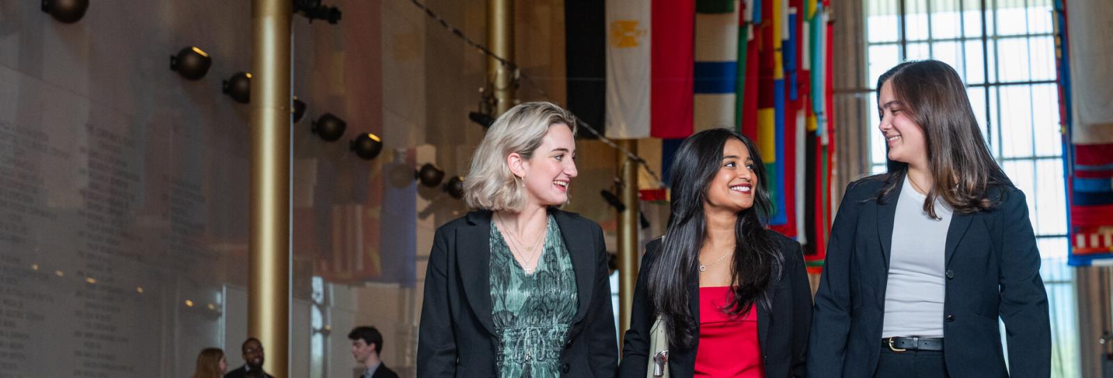 three business students walk through a lobby with many international flags hanging in the background