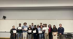 A group of students stands together indoors for a photo. They smile and hold certificates. 
