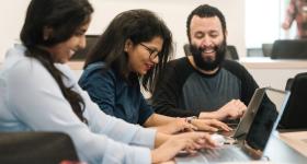 A group of three people sit and share a laugh. They look at laptops. 