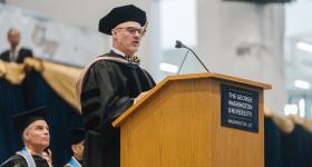 John Roberts, an alumnus of the GW School of Business speaks in front of a podium. He wears regalia.  Others can be seen  on the platform behind him. 