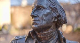 Image shows a broze statue of President George Washington with the GW University campus in the background.
