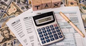 Image shows a desk with a calculator, tax forms, American currency, a pen, and a set of lettered wooden blocks that spell out the word "tax."