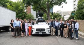 A large group of individuals dressed in professional attire stand in front of an autonomous vehicle. 