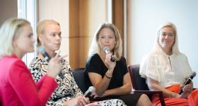 Three panelists listen as another speaks. All hold microphones. Each of the panel participants are women and appear to be seated in the front of a room and facing an audience.